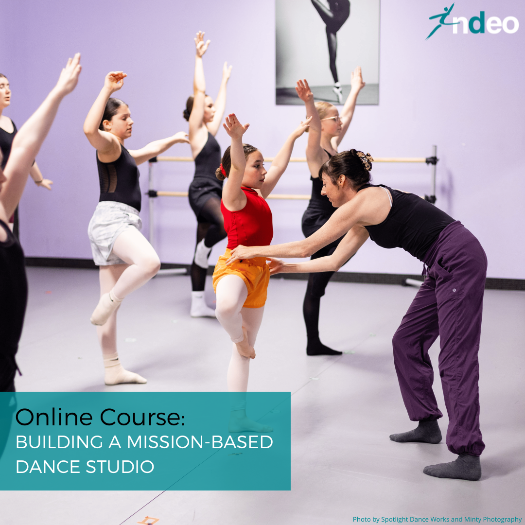 Dance teacher writing on board in studio with students in street clothes standing behind